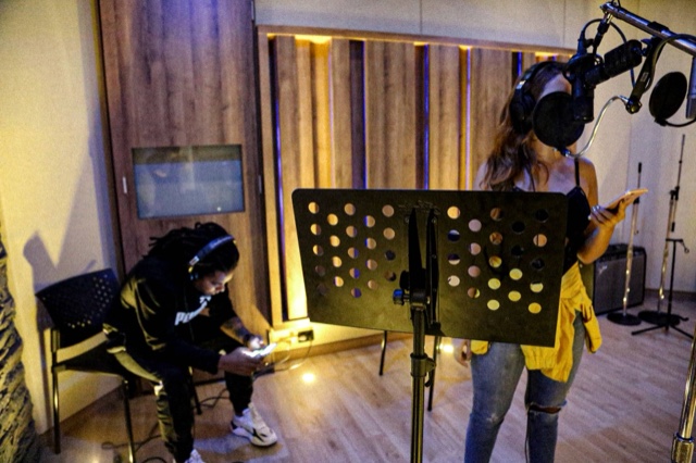 Colour photograph of a recording session in a studio in Medellín: Luigui Bleand sits with headphones on, looking at his phone, while Marilyn Oquendo records the song “La Movie” at a microphone, her face hidden by the pop filter